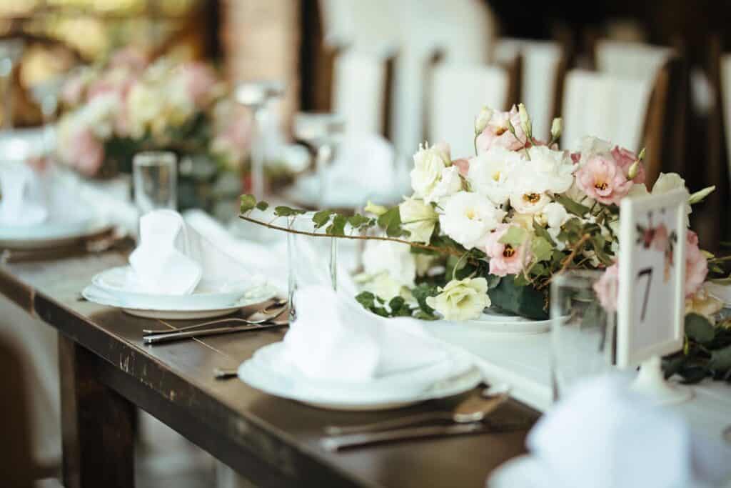 Lyst og moderne selskapslokale med stilren borddekking, hvite servietter og delikate blomster i pastellfarger – perfekt for bryllup eller konfirmasjon.